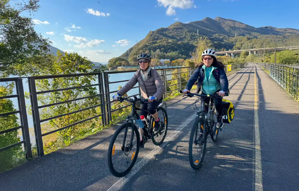 Two women riding road bikes along a riverside path in Korea on a sunny day during a long-distance cycling tour from Seoul to Andong